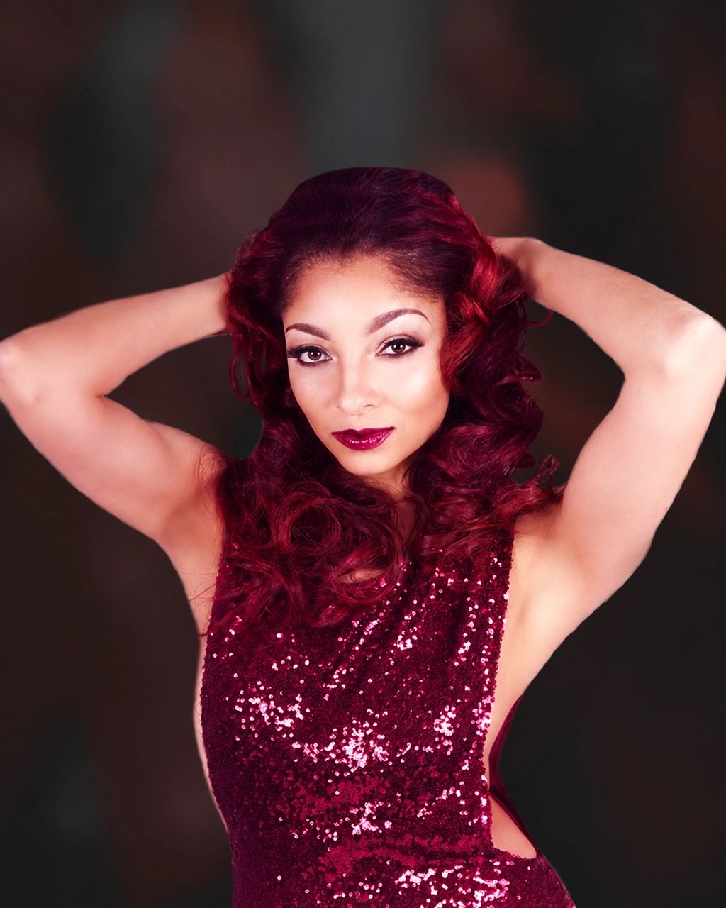 Woman with red curly hair in a burgundy sequin halter dress with both hands behind her head